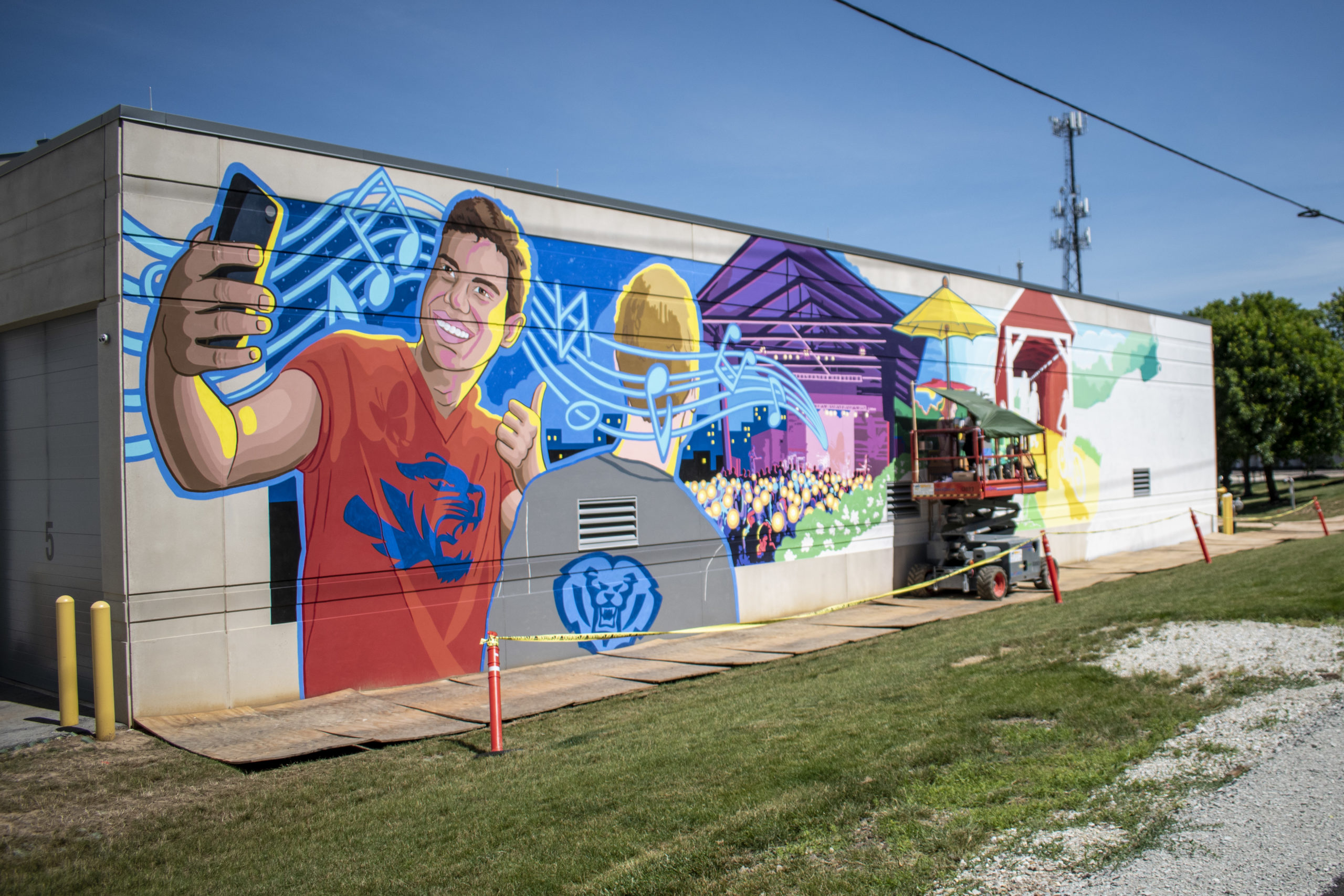 a mural of a person taking a selfie on their phone, a person with a school logo on their shirt, a concert venue with people and a covered bridge