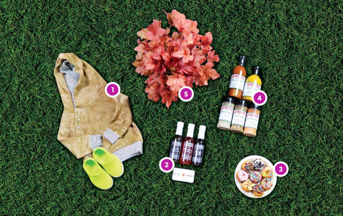 items laying on grass. starting from the far left is a brown sweatshirt with a white and black stripped inside. paired with the sweatshirt are green crocs. then a red and orange leaf plant. three bottles of a honey sauce. two bottles of bbq sauce and underneath that are three bottles of meat rub and seasonings. then a small plate with an assortment of miniature donuts.