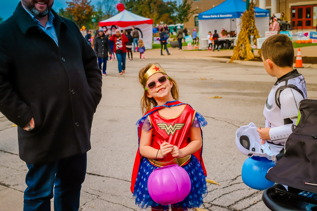 a girl smiling in a wonder woman costume while holding a pink trick or treat pumpkin bucket